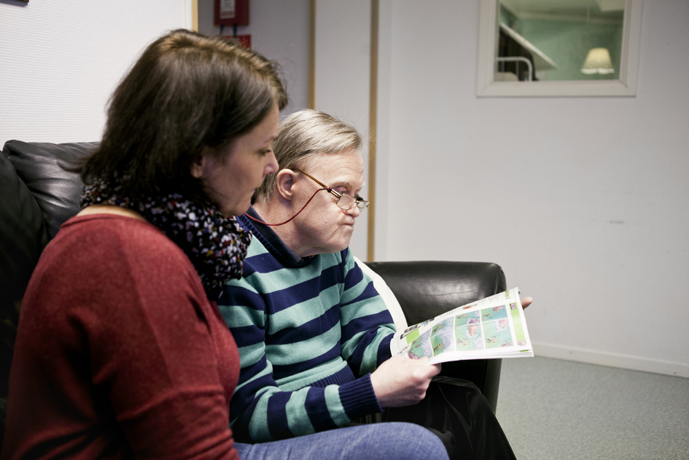 Caregiver looking at mentally challenged senior man reading magazine at home